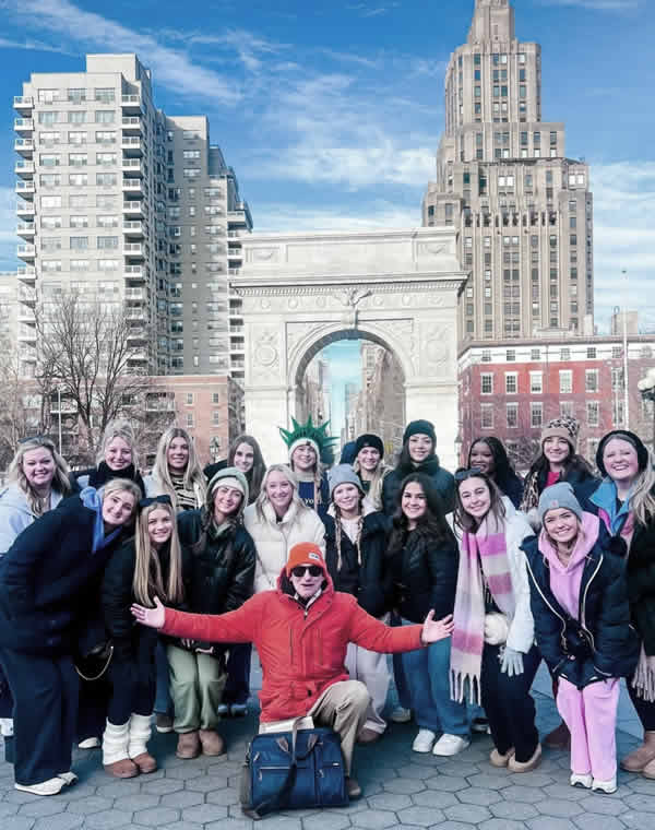 Jared Goldstein in an orange jacket kneeling in front of a large group of his tourists in WAshington Square Park in front of the Washington Share Park Arch